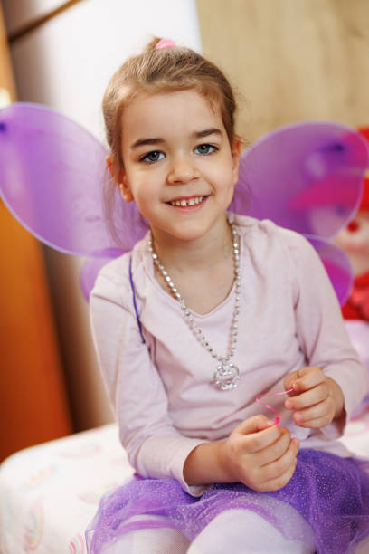 Portrait of a cute happy little girl in purple fairy costume, sitting on bed and looking at camera at home